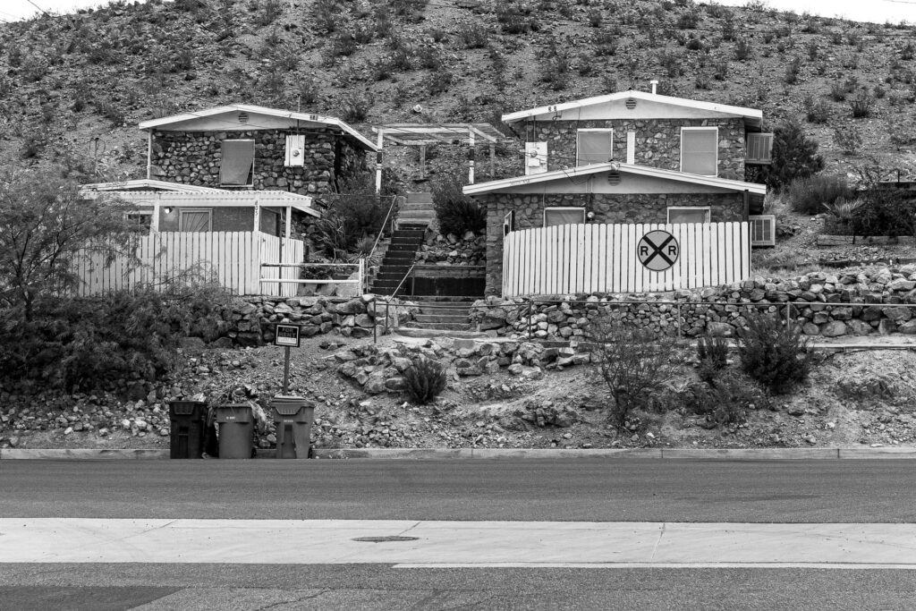 Desert stone houses on a rocky hillside with a "For Rent" sign, trash bins by the street, and a yellow railroad crossing symbol on a white fence.