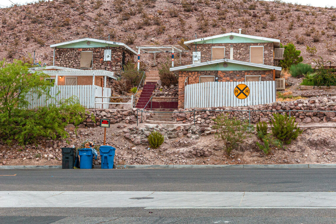 Desert stone houses on a rocky hillside with a "For Rent" sign, trash bins by the street, and a yellow railroad crossing symbol on a white fence.