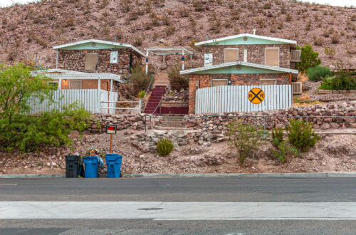 Desert stone houses on a rocky hillside with a "For Rent" sign, trash bins by the street, and a yellow railroad crossing symbol on a white fence.