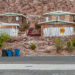 Desert stone houses on a rocky hillside with a "For Rent" sign, trash bins by the street, and a yellow railroad crossing symbol on a white fence.
