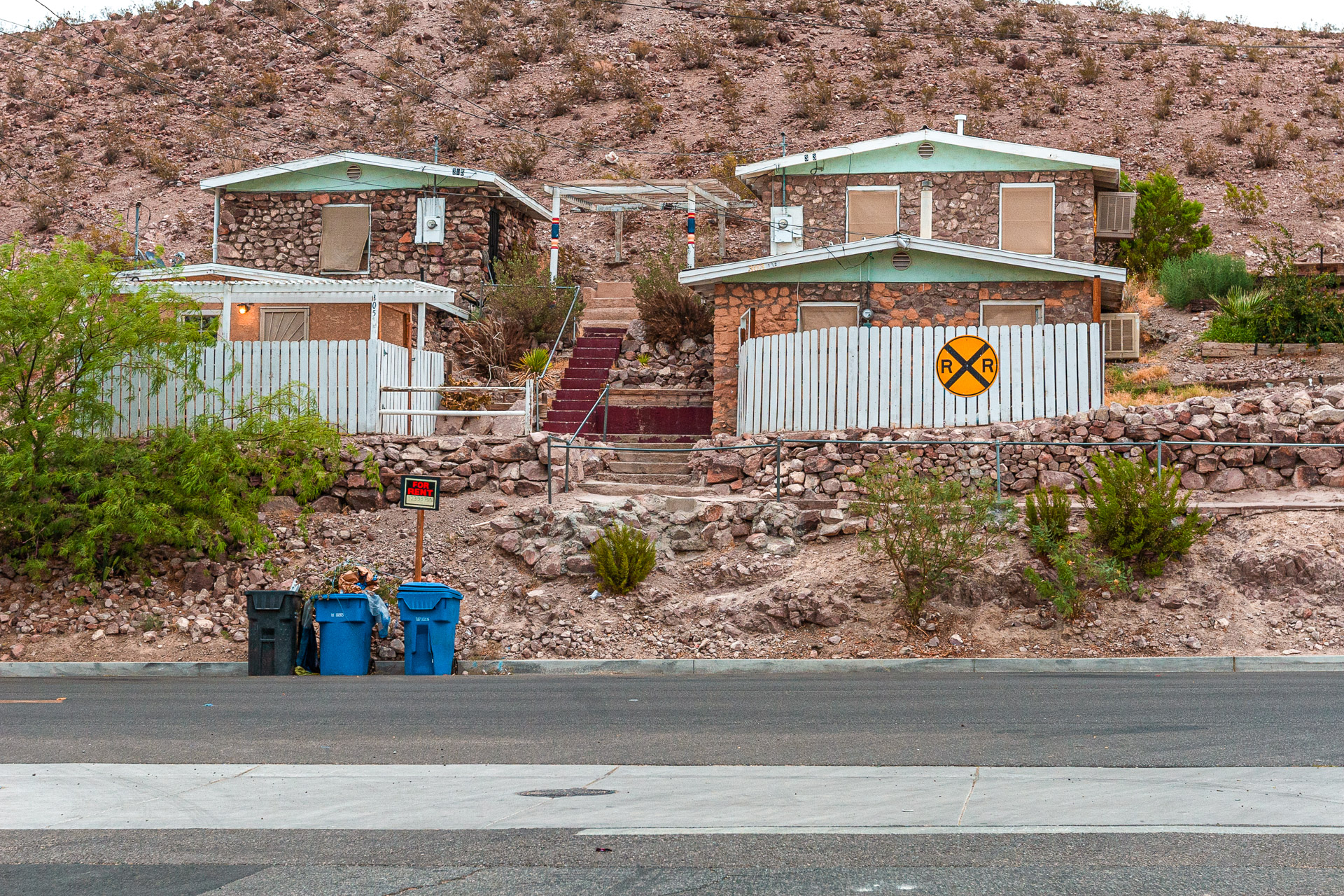 Desert stone houses on a rocky hillside with a "For Rent" sign, trash bins by the street, and a yellow railroad crossing symbol on a white fence.