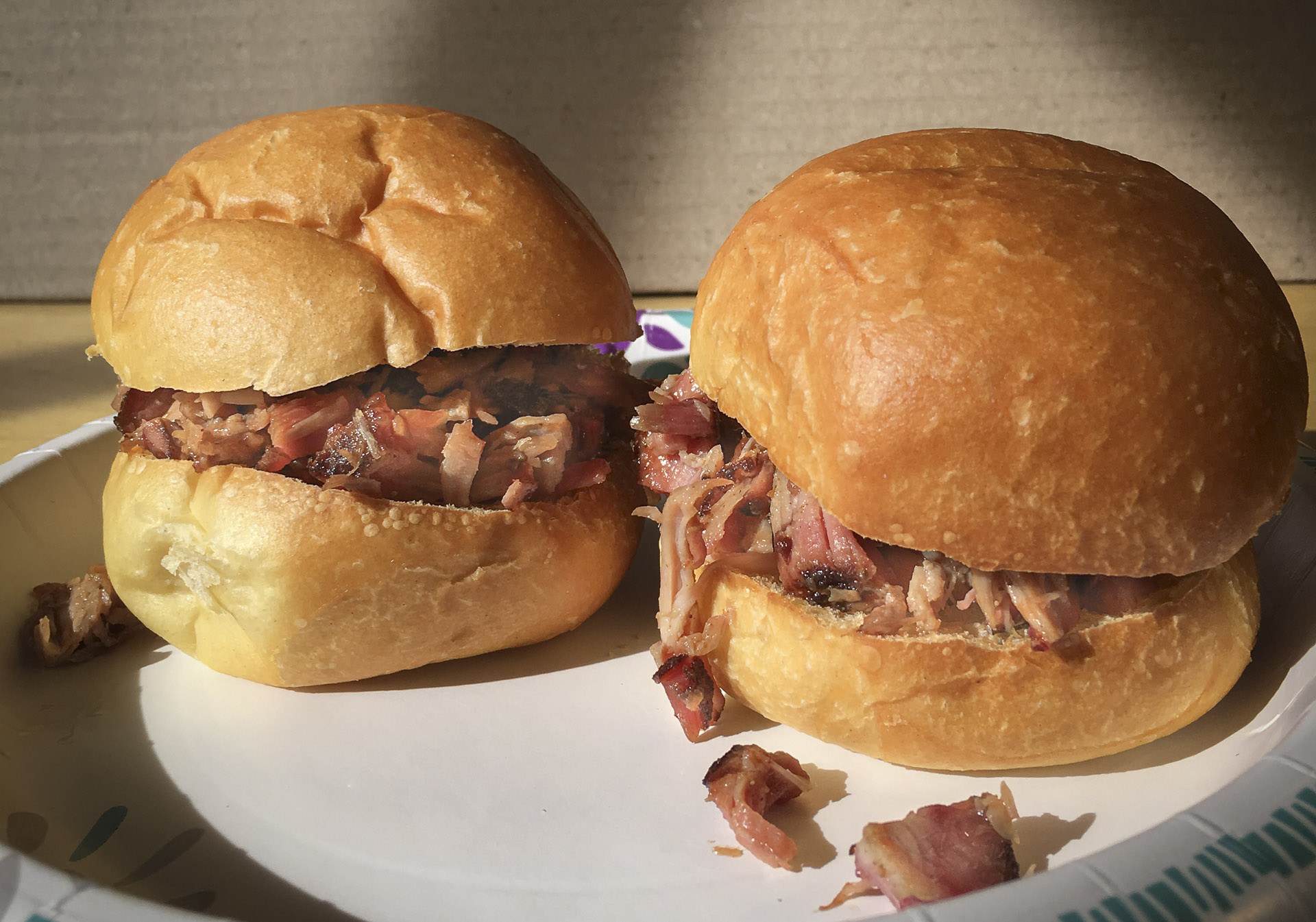 Two pulled‑pork sandwiches on a white paper plate, each with a soft golden bun and shredded smoked pork spilling slightly onto the plate.