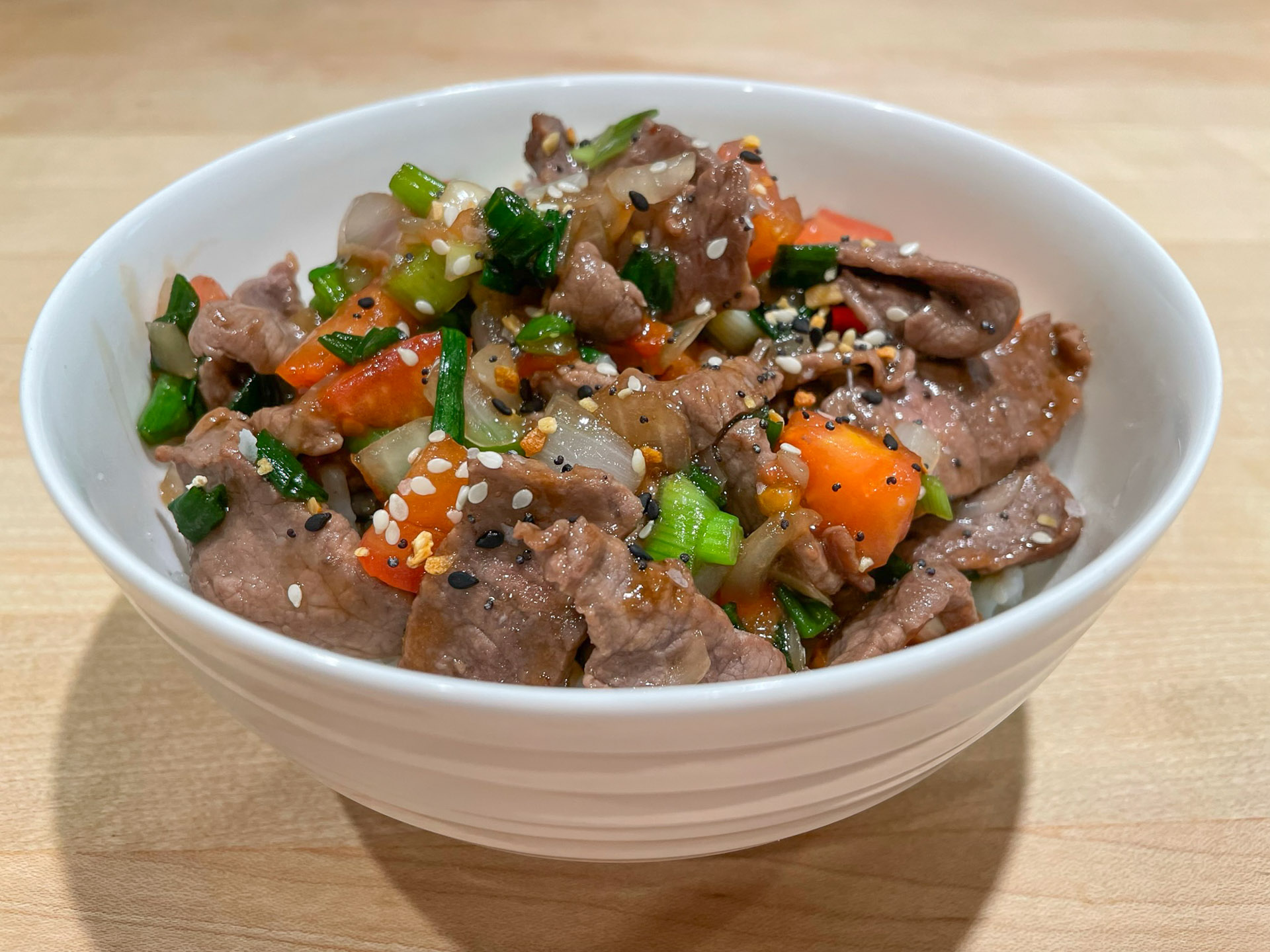 Close‑up of a beef stir‑fry bowl with thinly sliced beef, colorful bell peppers, green onions, and sesame seeds served in a white ceramic bowl on a wooden surface.