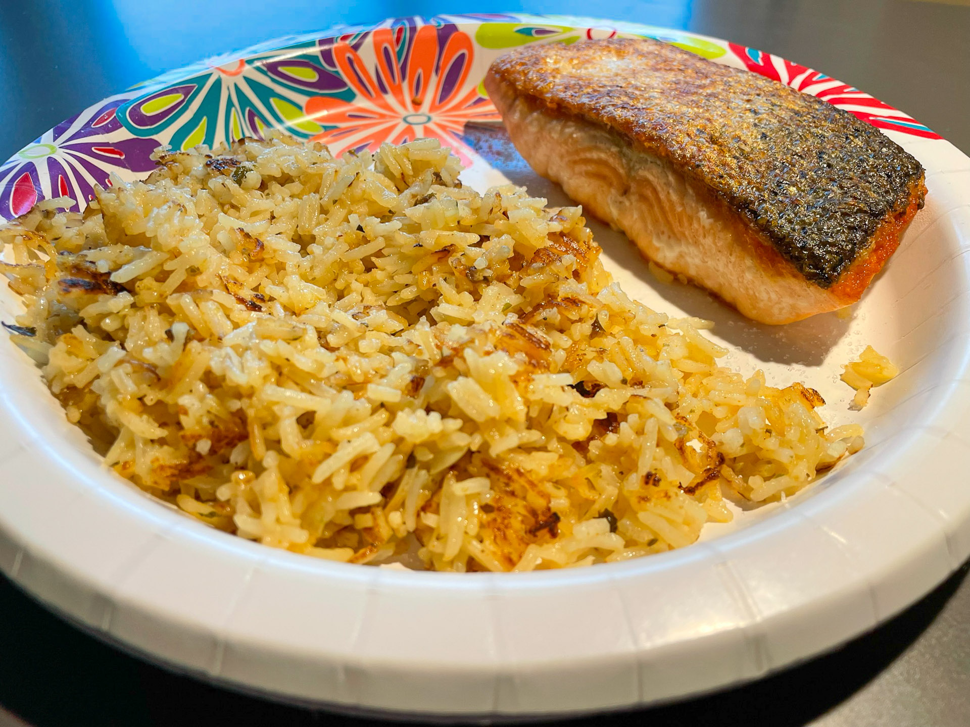 Crispy‑skinned pan‑seared salmon fillet served with golden fried rice on a colorful floral paper plate, photographed under natural light for a home‑cooked meal presentation.