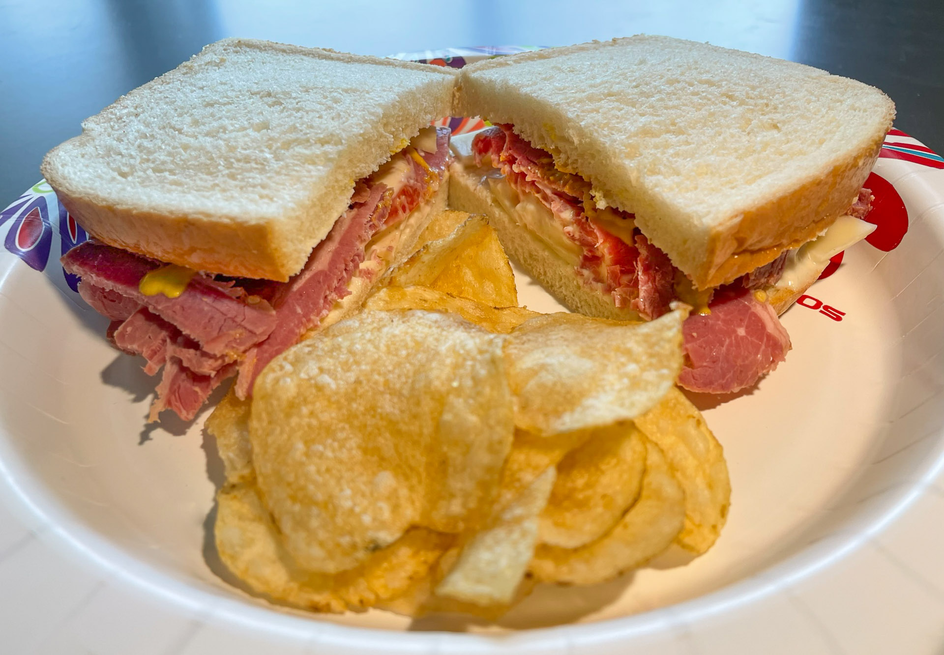 Close‑up of a pastrami sandwich with mustard and cheese on white bread, served with crispy potato chips on a colorful paper plate.