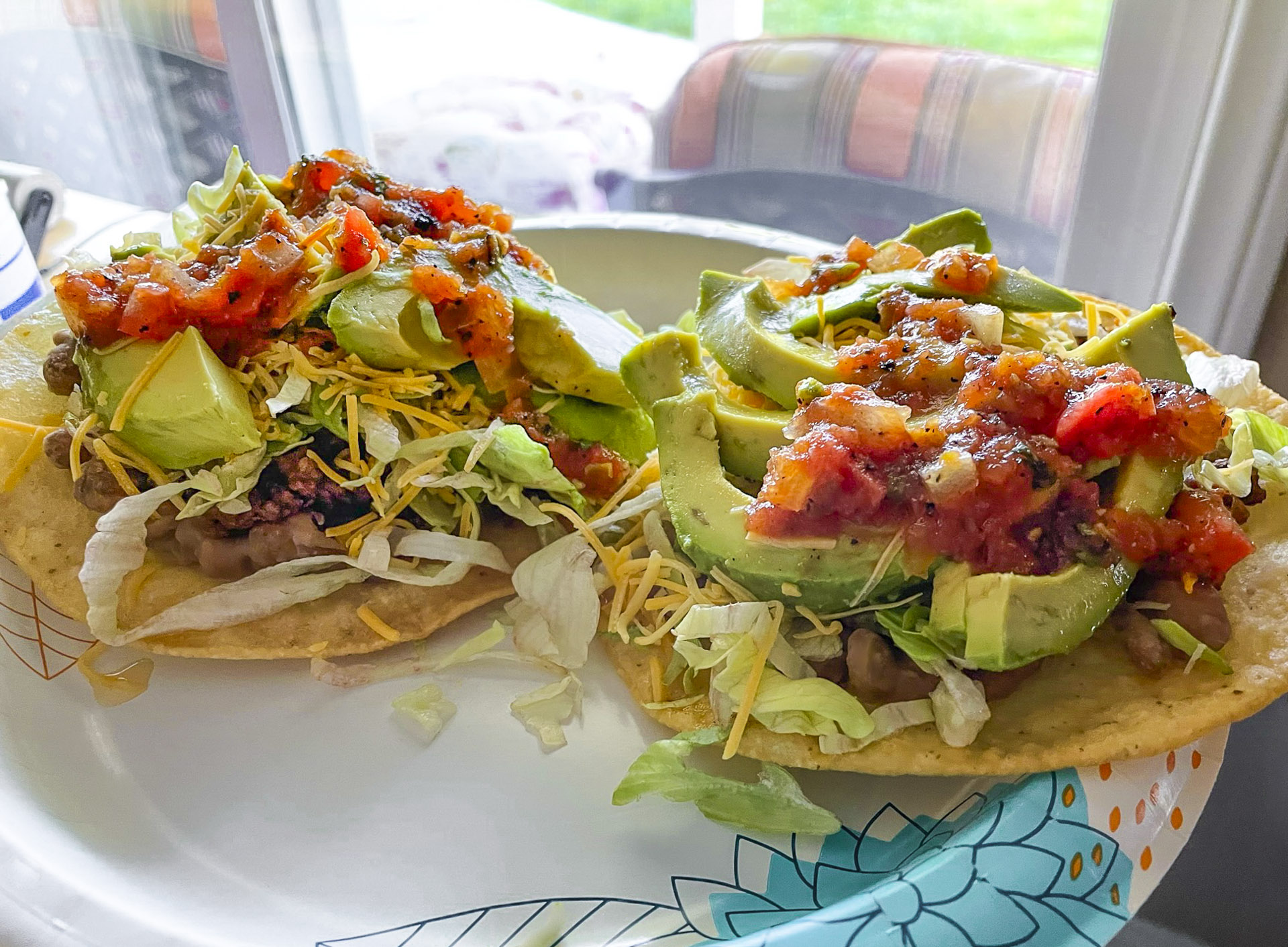 Two loaded Mexican tostadas on a patterned paper plate, topped with refried beans, shredded lettuce, shredded cheese, thick slices of fresh green avocado, and chunky red tomato salsa.