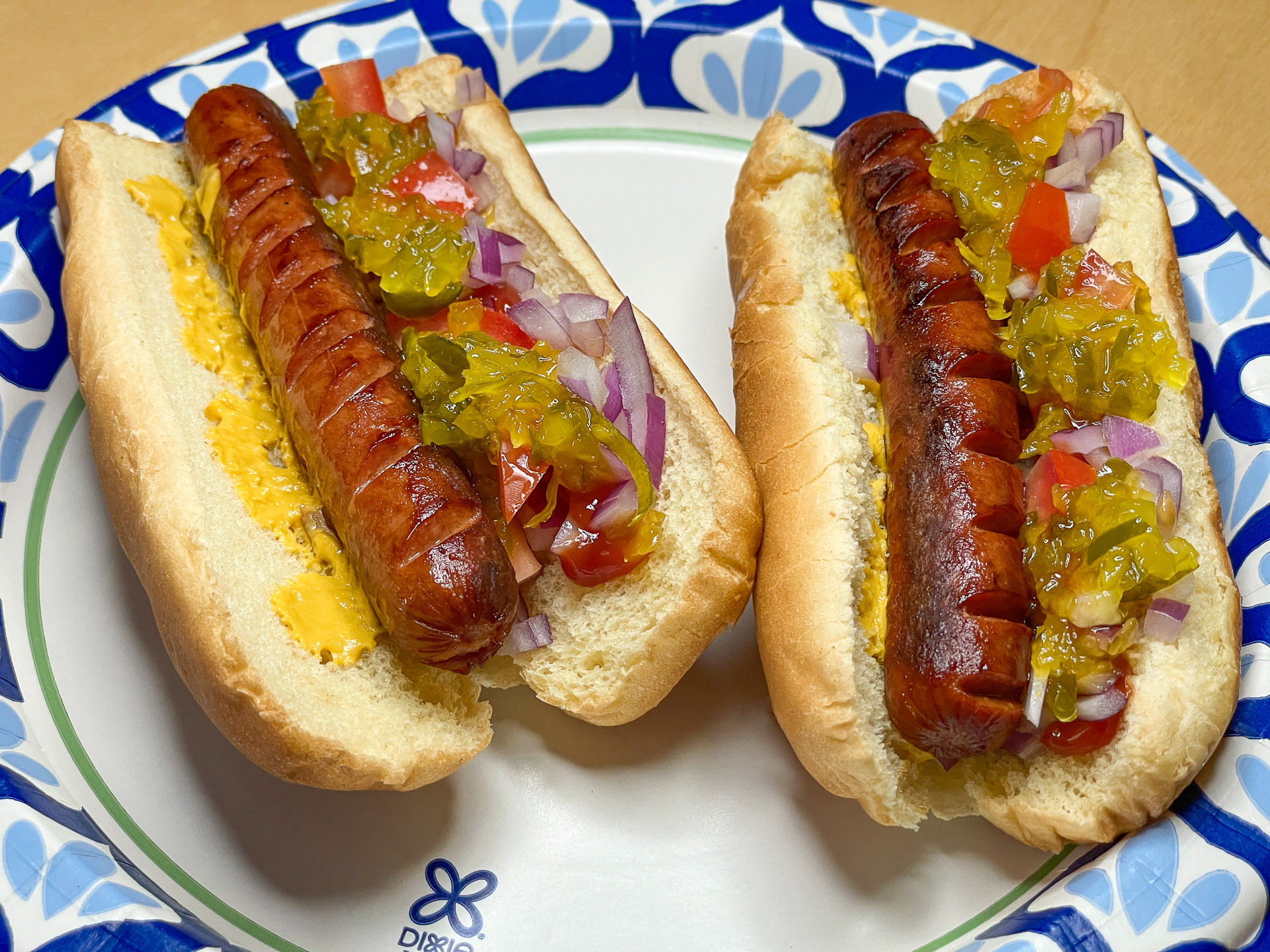A pair of grilled hot dogs in buns topped with mustard, diced tomatoes, chopped red onions, and green relish on a blue‑and‑white floral paper plate.