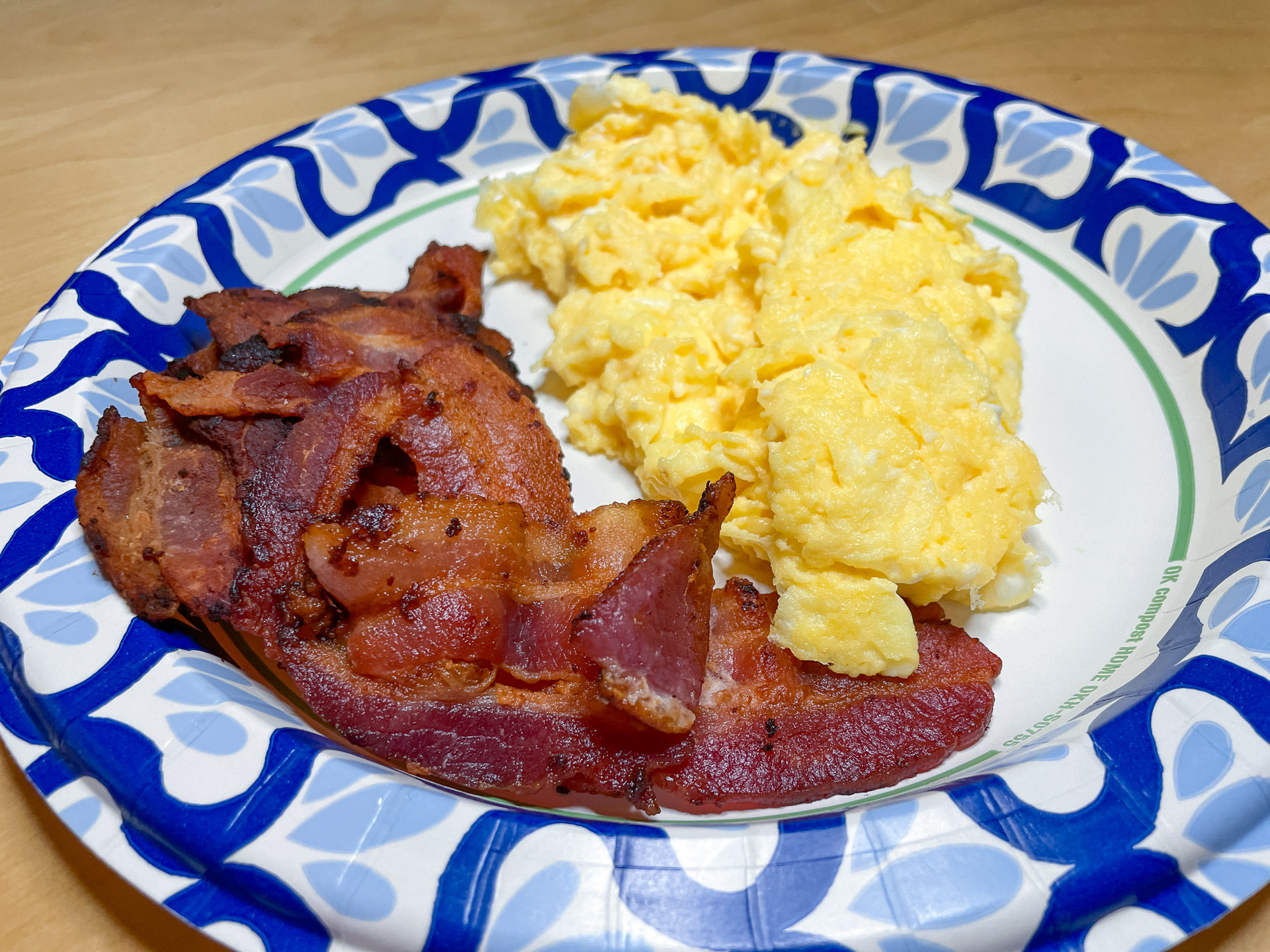 A breakfast plate featuring a pile of fluffy scrambled eggs and several strips of crispy, cooked bacon served on a blue and white patterned paper plate.