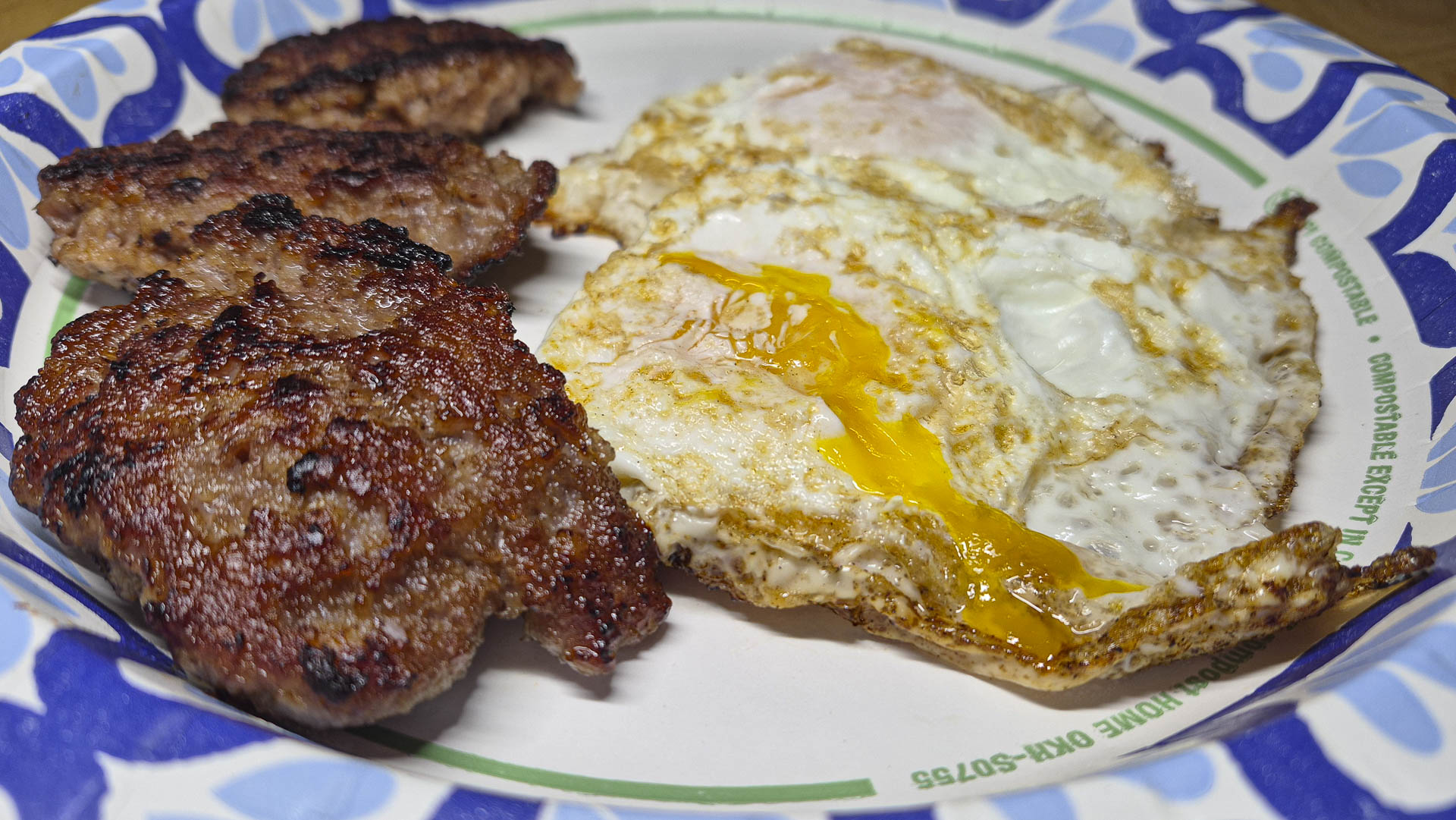 Close‑up of a hearty breakfast plate with three browned sausage patties and two fried eggs, one with a broken yolk, served on a blue‑and‑white paper plate.