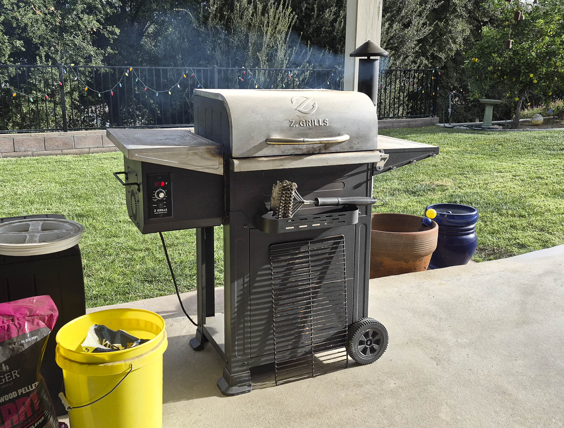 Outdoor pellet grill with smoke rising from the lid, featuring a Z Grills logo, control panel, and barbecue tools on a sunny backyard patio surrounded by green grass and garden pots.
