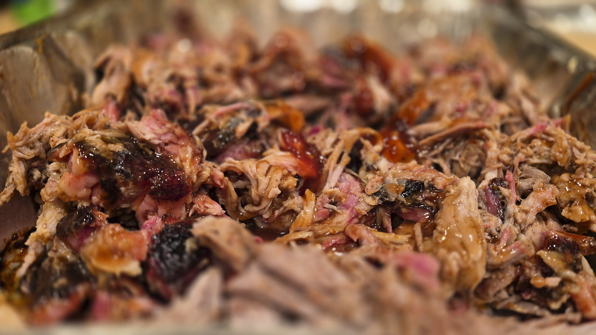 Close‑up of tender pulled pork with smoky bark and glistening barbecue sauce, showing juicy shredded meat texture and caramelized edges.