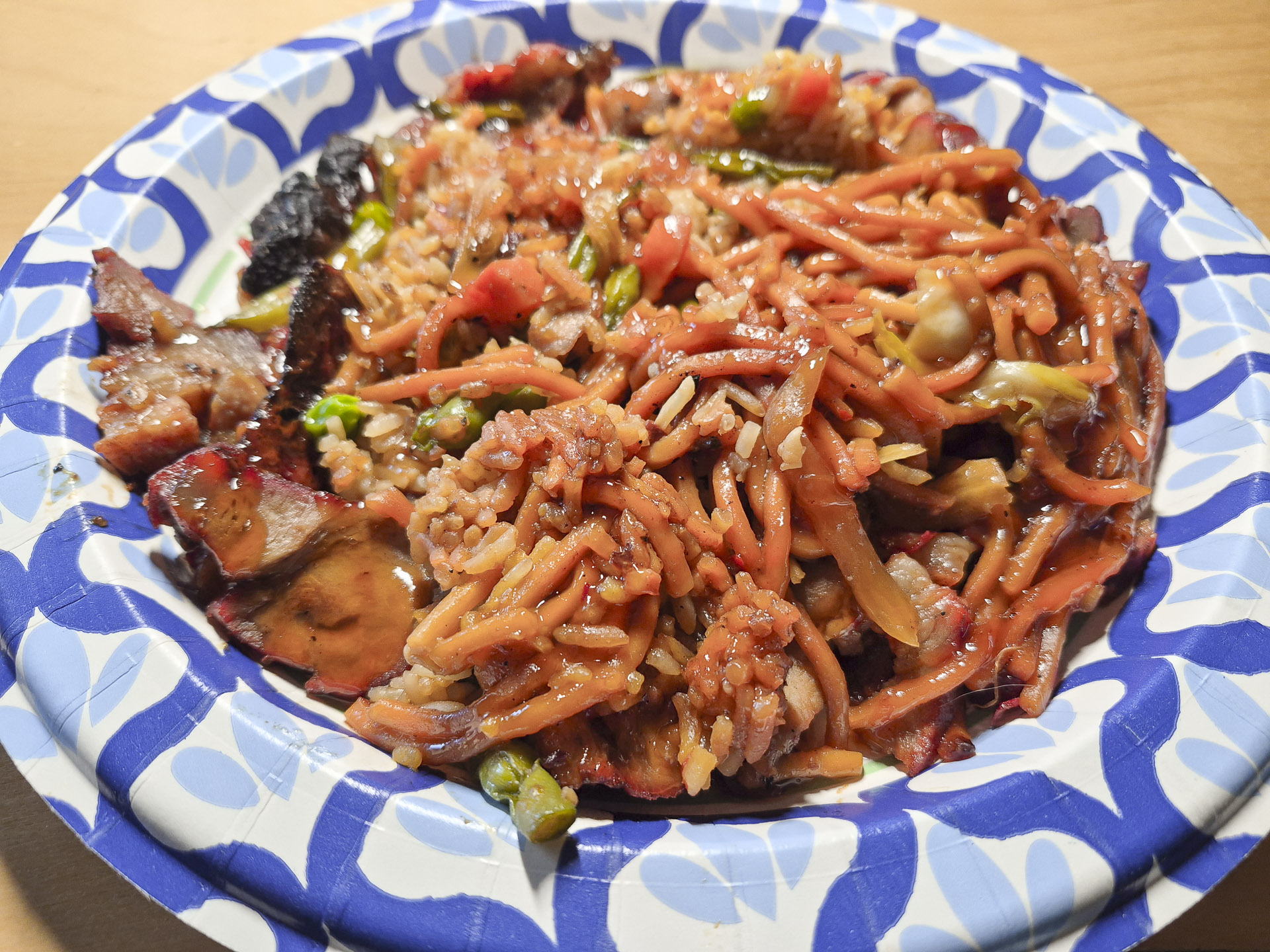 Close‑up of pork chow mein with rice, vegetables, and savory sauce served on a decorative paper plate, featuring noodles, peas, carrots, and char‑siu pork slices.