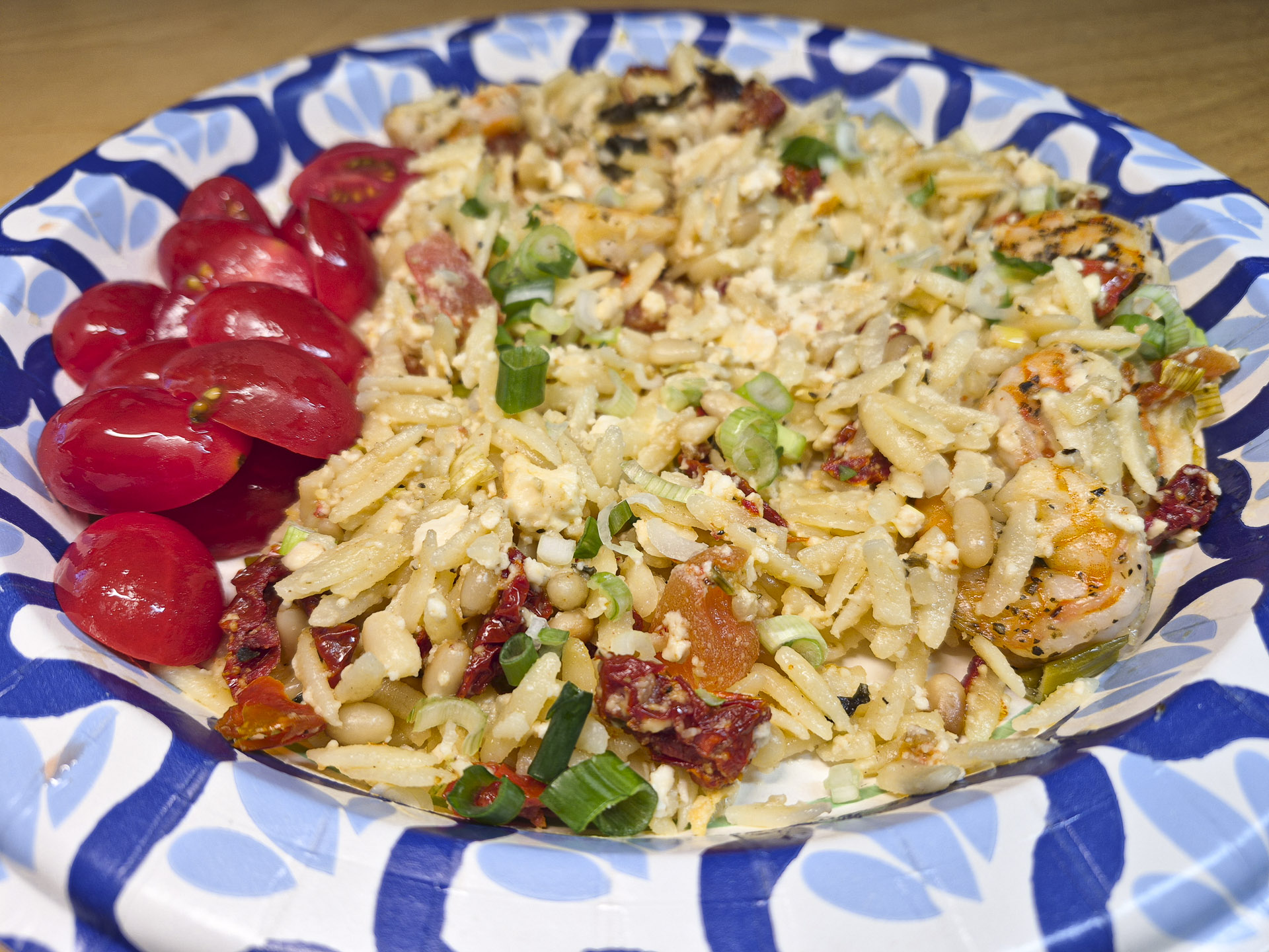 Close‑up of Mediterranean shrimp orzo salad with feta cheese, sun‑dried tomatoes, pine nuts, and sliced grape tomatoes served on a decorative paper plate.