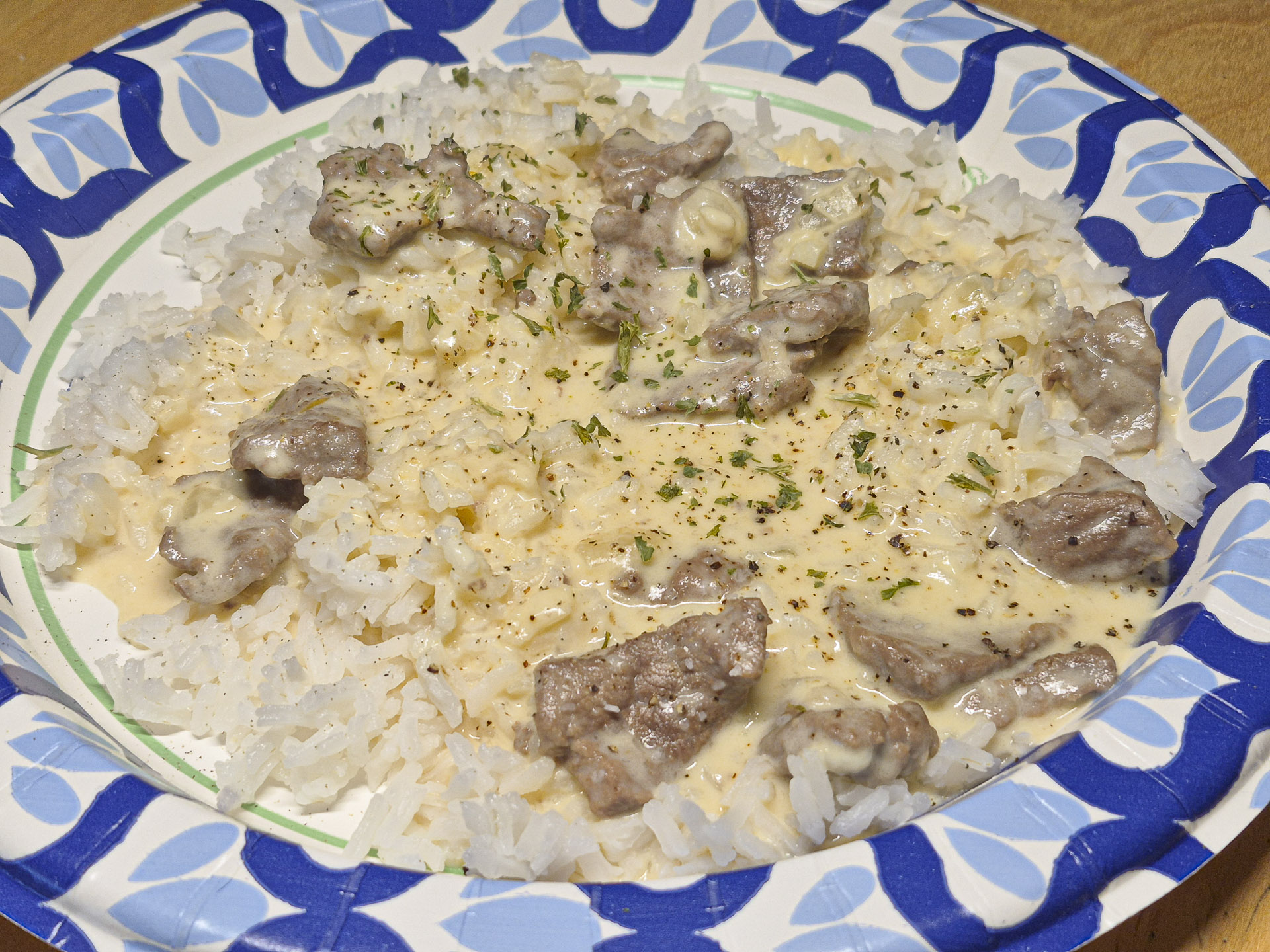 Close‑up of beef stroganoff and rice dish served on a decorative paper plate, featuring tender beef pieces in a rich cream sauce with herbs and black pepper garnish.