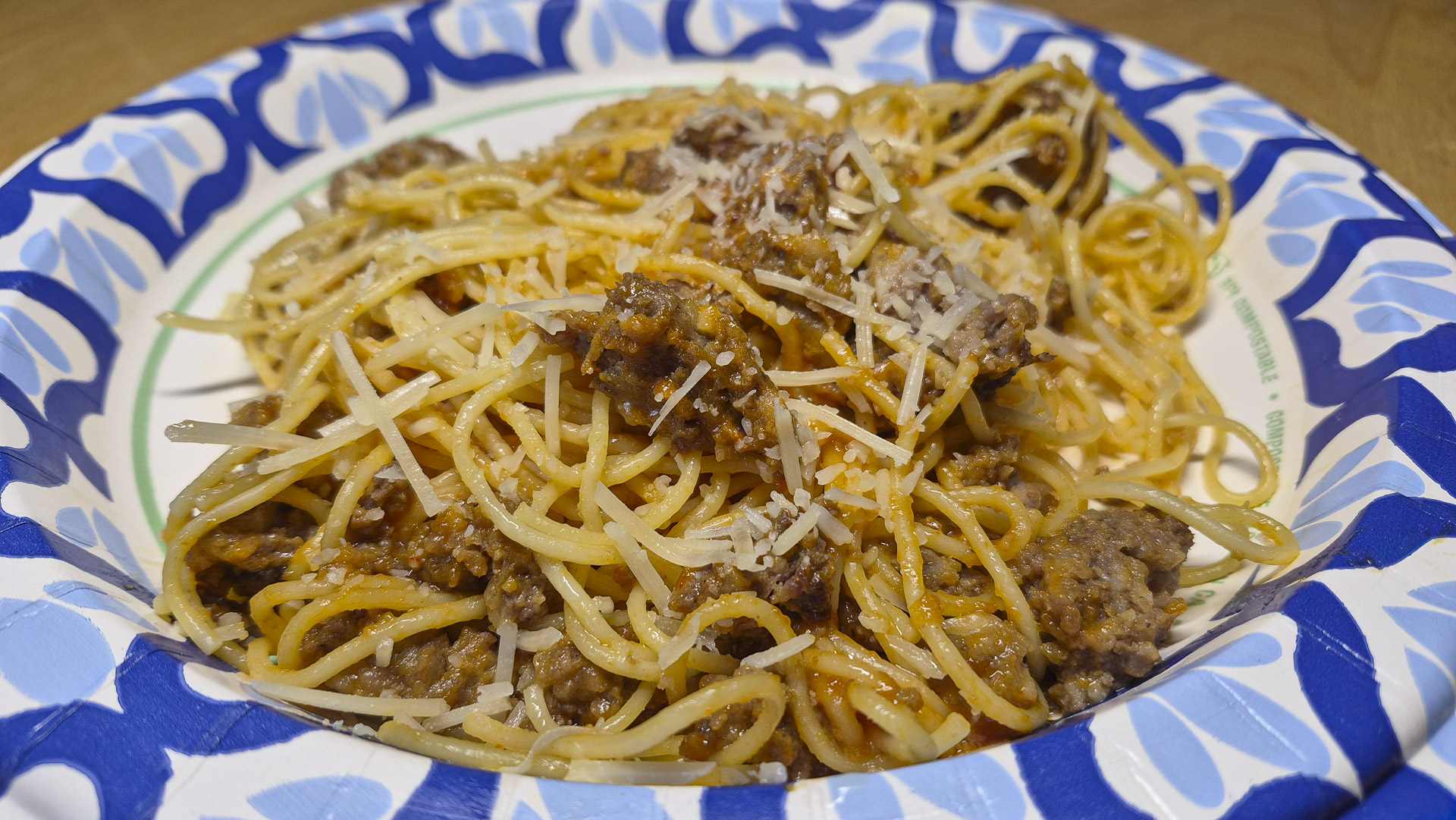 Close‑up of spaghetti with meat sauce and grated Parmesan cheese served on a decorative paper plate, showing golden pasta strands and savory beef sauce texture.