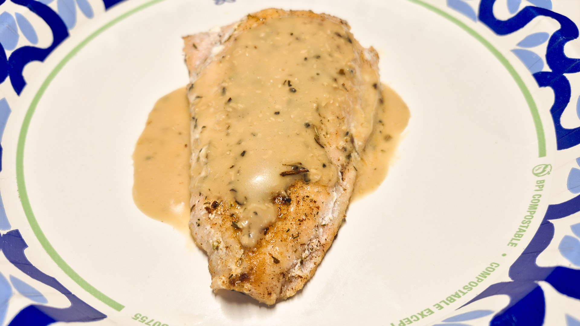 Close‑up of a seasoned fish fillet topped with creamy herb sauce on a decorative paper plate, highlighting golden‑brown edges and smooth texture.
