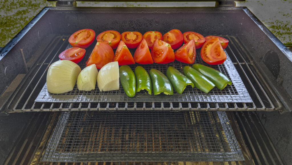 Fresh vegetables including halved tomatoes, quartered onions, and jalapeƱo peppers arranged neatly on a grill rack, ready for roasting