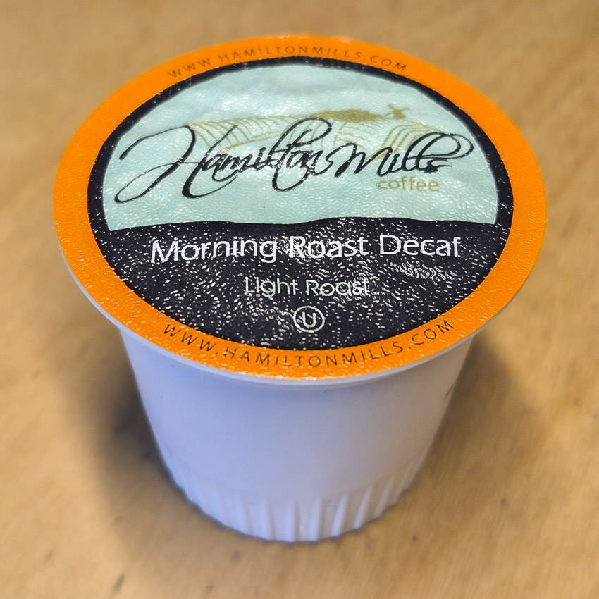 Hamilton Mills Morning Roast Decaf coffee pod with orange rim and branded lid on a wooden surface.