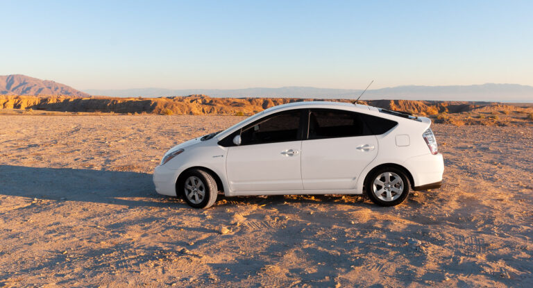 White Toyota Prius parked on a sandy desert overlook, with sunlit rocky ridges and distant mountains under a clear sky.