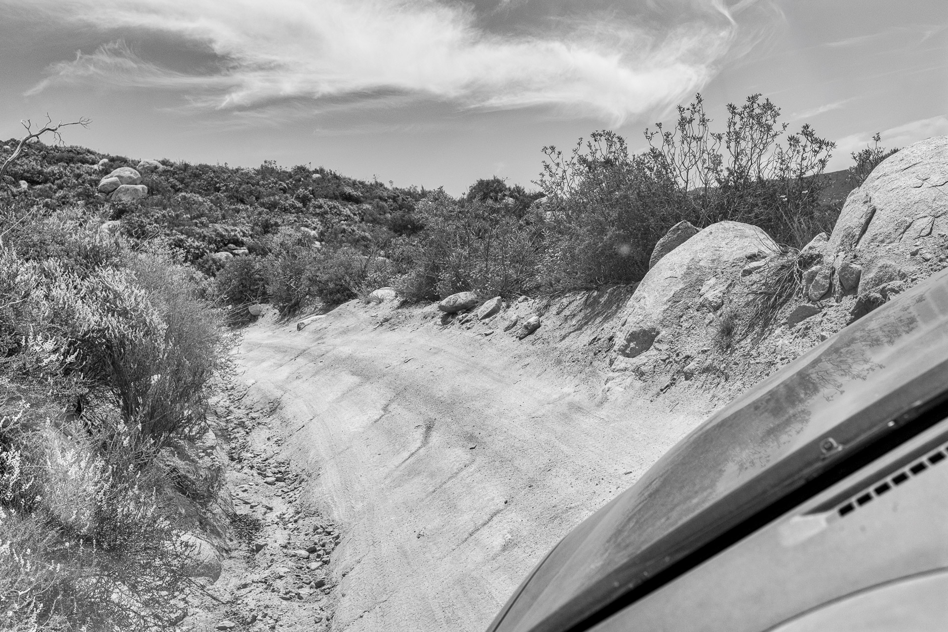 Black-and-white photograph of a rugged off-road trail winding through rocky desert terrain, with scattered boulders, low shrubs, dramatic clouds, and part of a vehicle visible in the foreground.