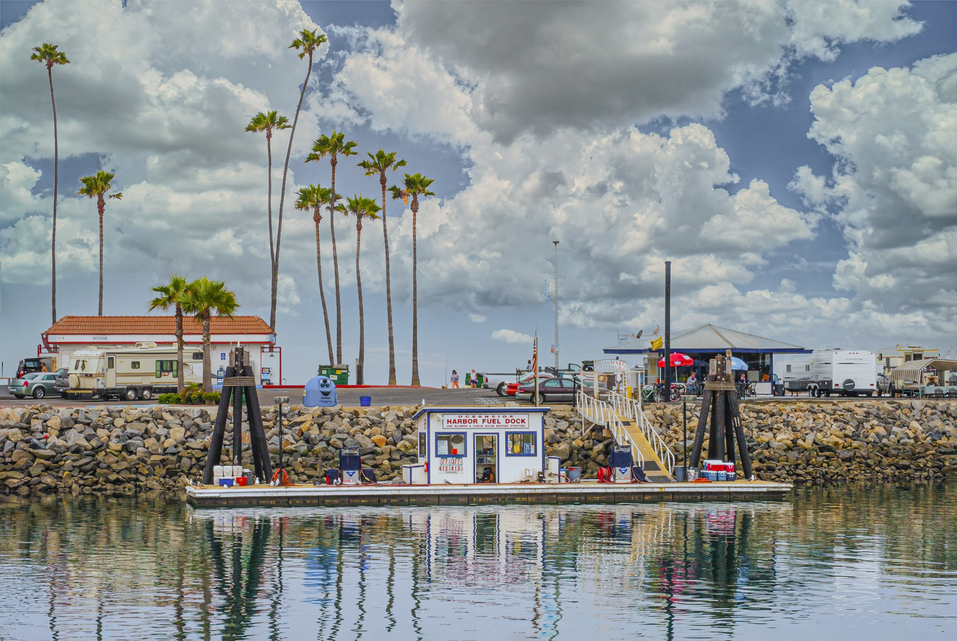 Floating harbor fuel dock in Oceanside, California, with palm trees, boats, and dramatic clouds overhead.