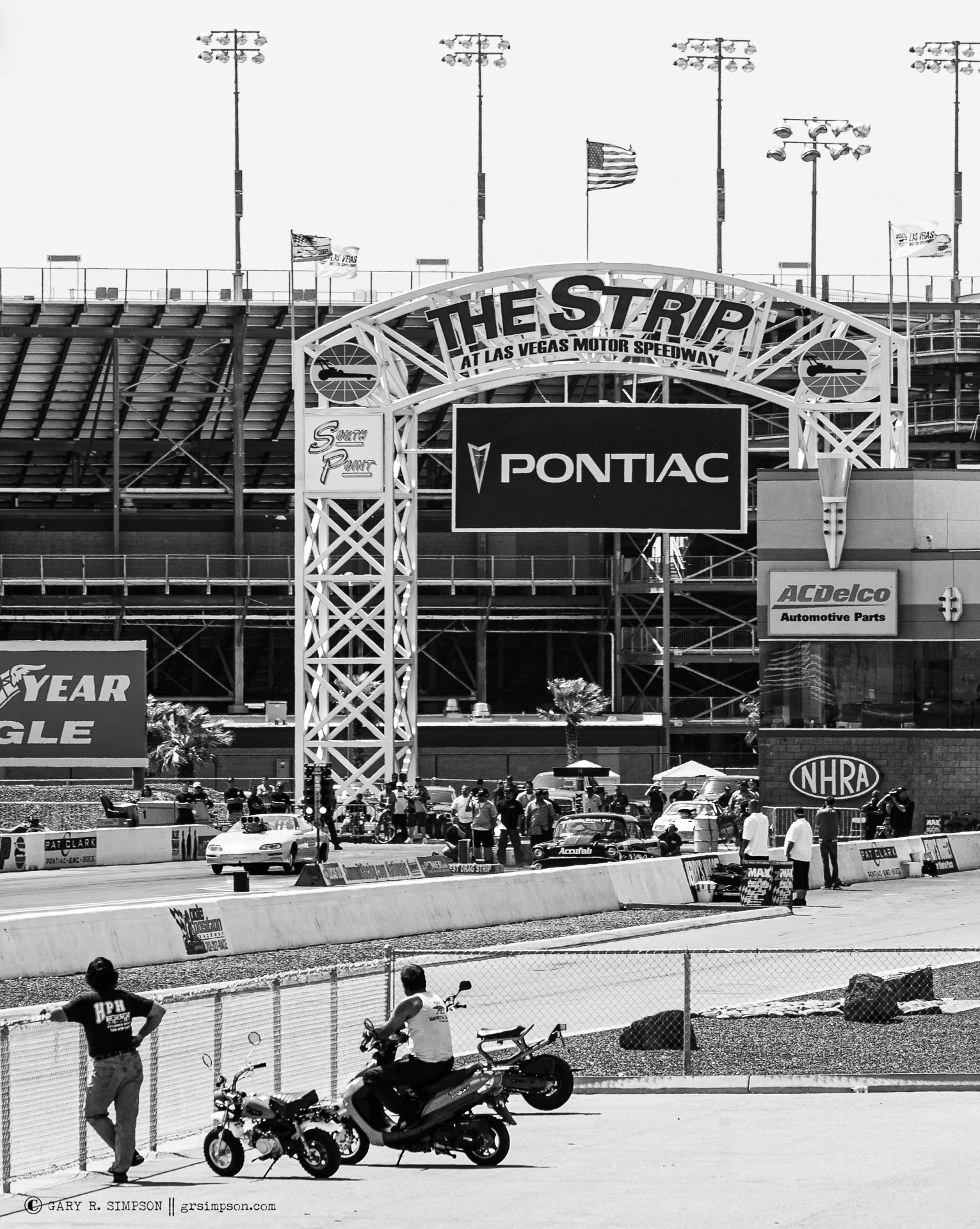 Black-and-white photograph of drag racing cars beneath The Strip sign at Las Vegas Motor Speedway.