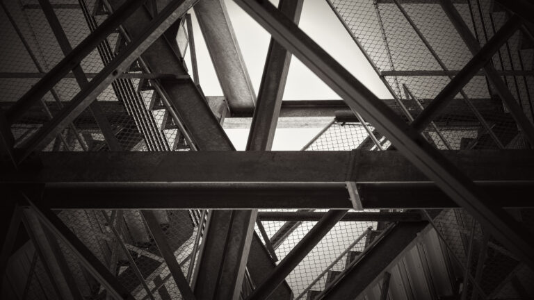Monochrome view looking up beneath bleachers, with intersecting steel beams and wire mesh framing angular patches of bright sky.