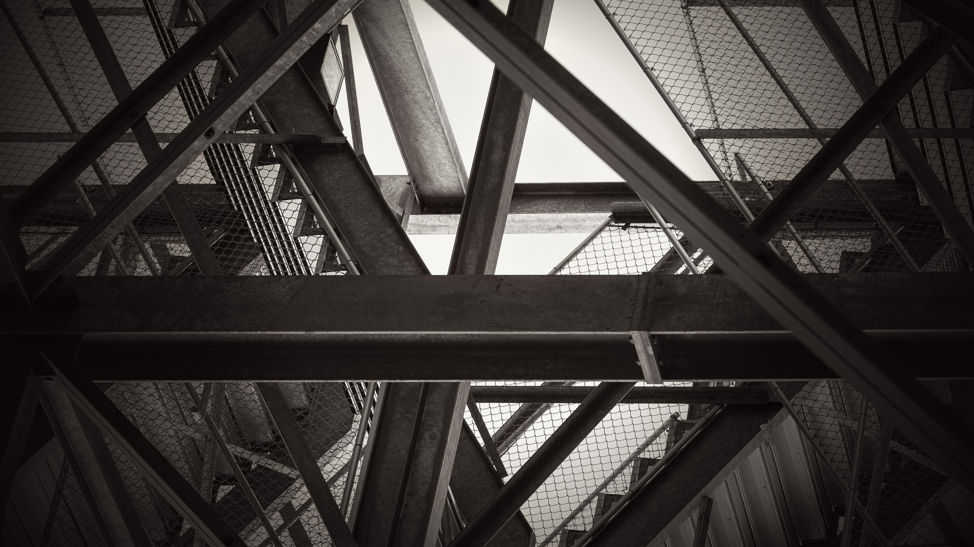 Monochrome view looking up beneath bleachers, with intersecting steel beams and wire mesh framing angular patches of bright sky.