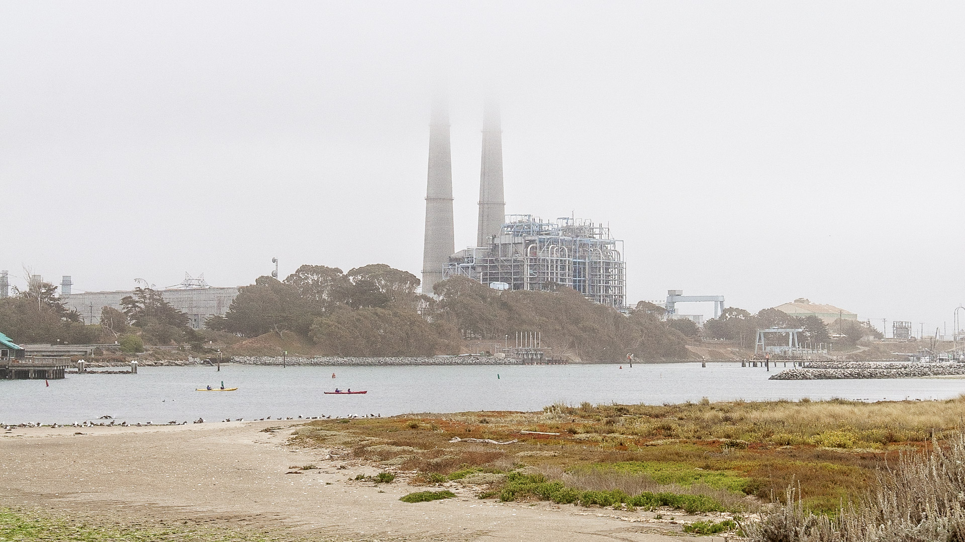 Moss Landing Power Plant and its twin smokestacks rising through coastal fog above the harbor, with kayakers on the water.