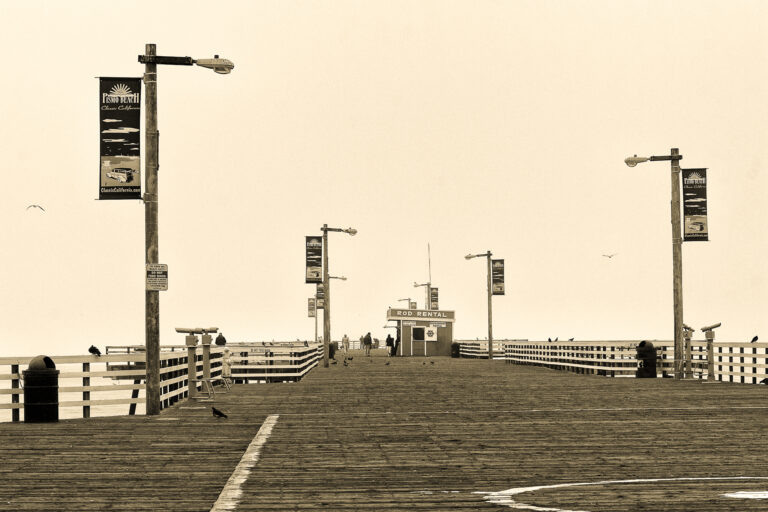 Sepia-toned view along the wooden Pismo Beach Pier, with railings, lampposts, banners, a fishing rod rental booth, and a few people and birds in the distance.