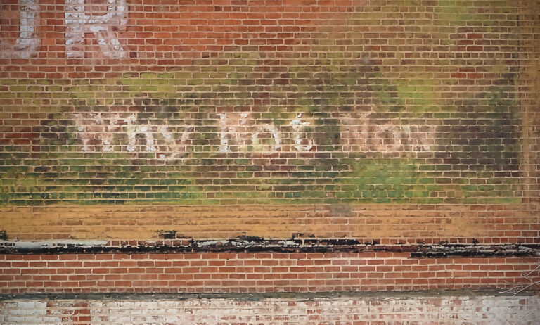 Faded early advertising painted on a weathered brick wall, with remnants of white lettering and layers of red, green, and yellow paint.