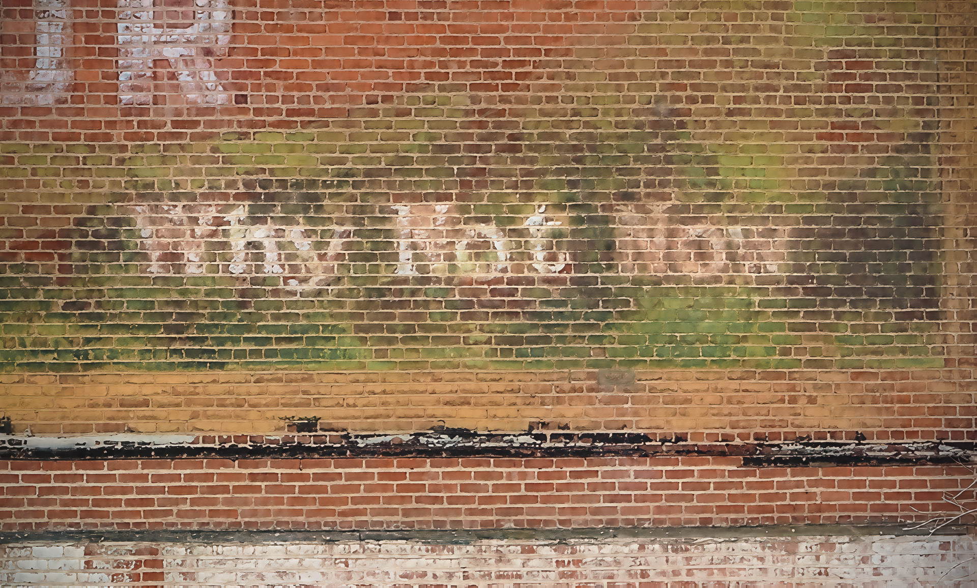 Faded early advertising painted on a weathered brick wall, with remnants of white lettering and layers of red, green, and yellow paint.