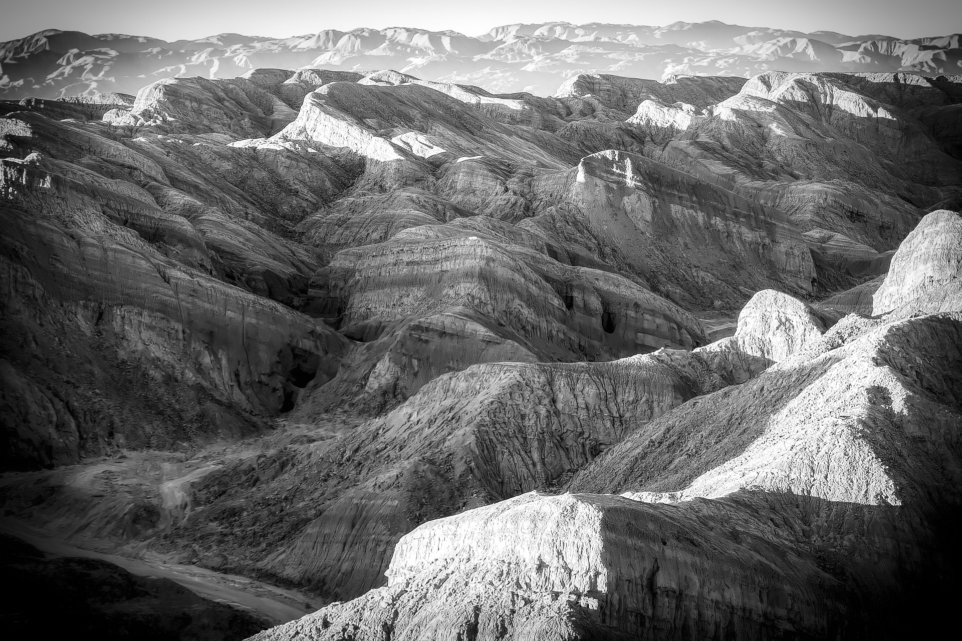 Black-and-white landscape photograph of rugged, eroded badlands with rounded hills, deep gullies, and layered rock formations illuminated by contrasting sunlight and shadow.