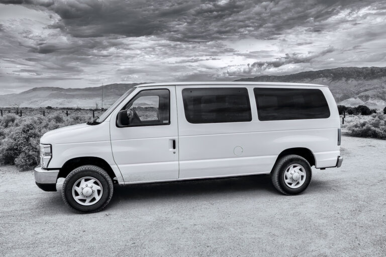 Black-and-white side view of a white Ford passenger van parked on a desert roadside, with scrub vegetation, distant mountains, and heavily clouded skies.