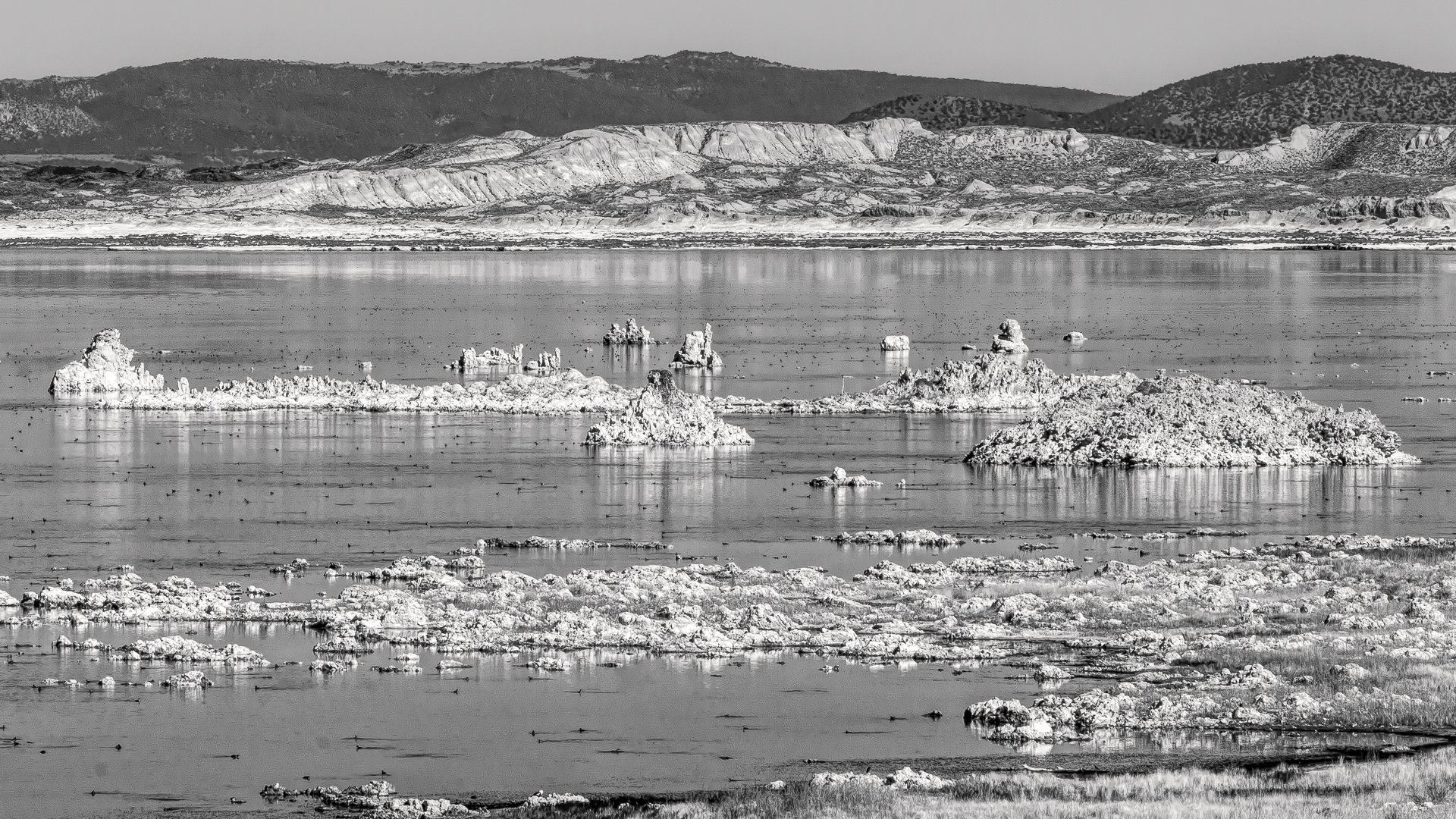 Black-and-white landscape photograph of tufa rock formations rising from a calm lake, with rugged hills and pale rocky slopes in the background.