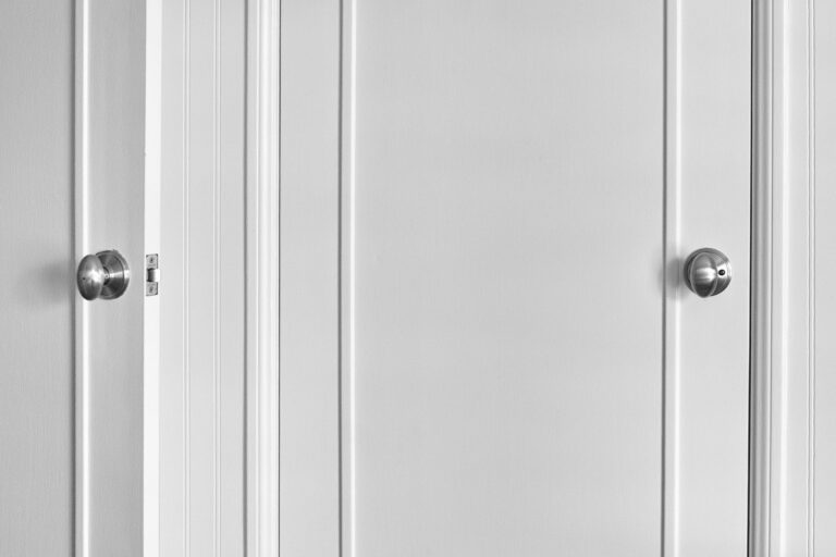 Black-and-white photograph of two white doors with silver doorknobs, one partially open and the other closed, separated by vertical wall panels.
