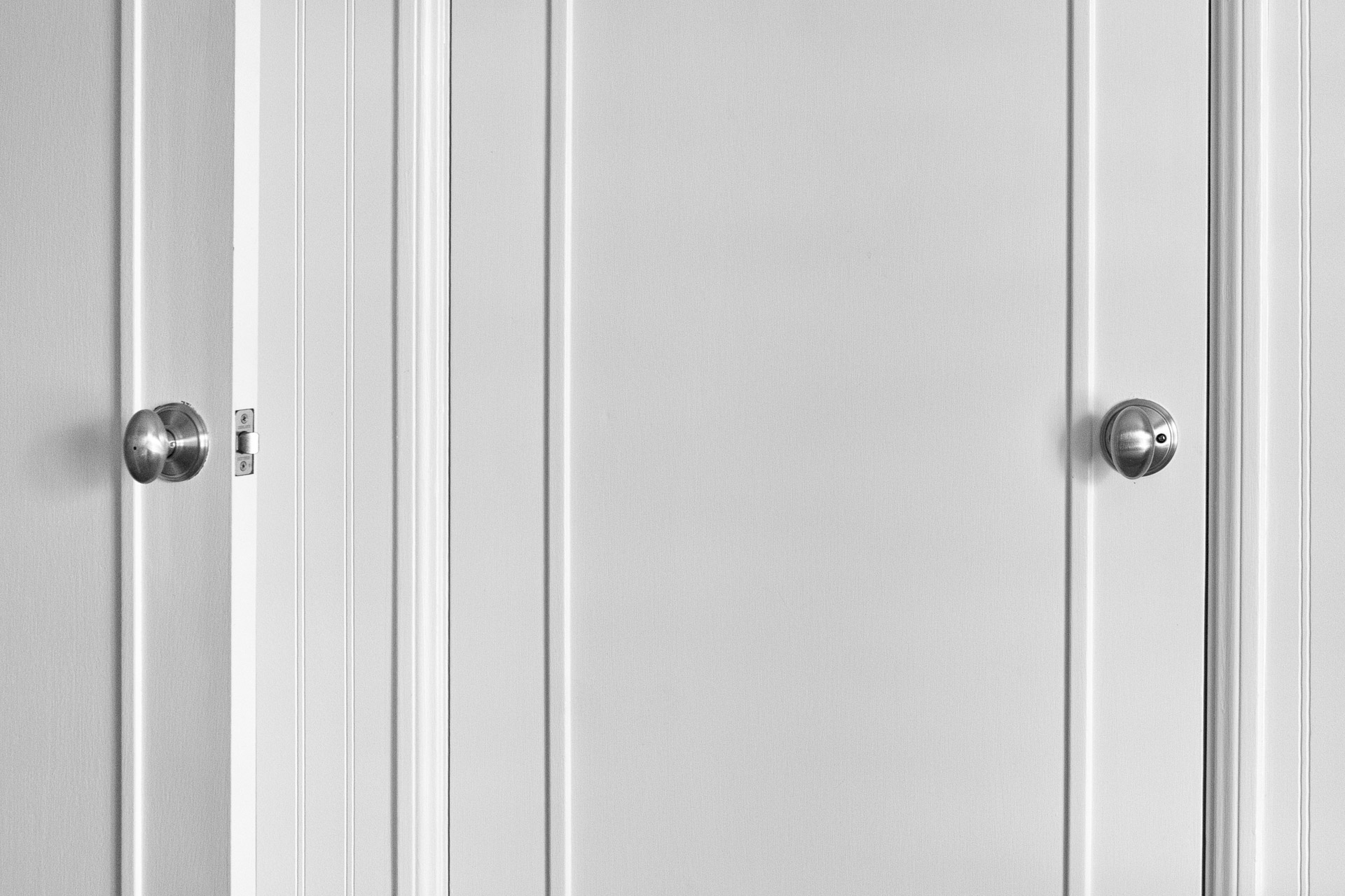 Black-and-white photograph of two white doors with silver doorknobs, one partially open and the other closed, separated by vertical wall panels.
