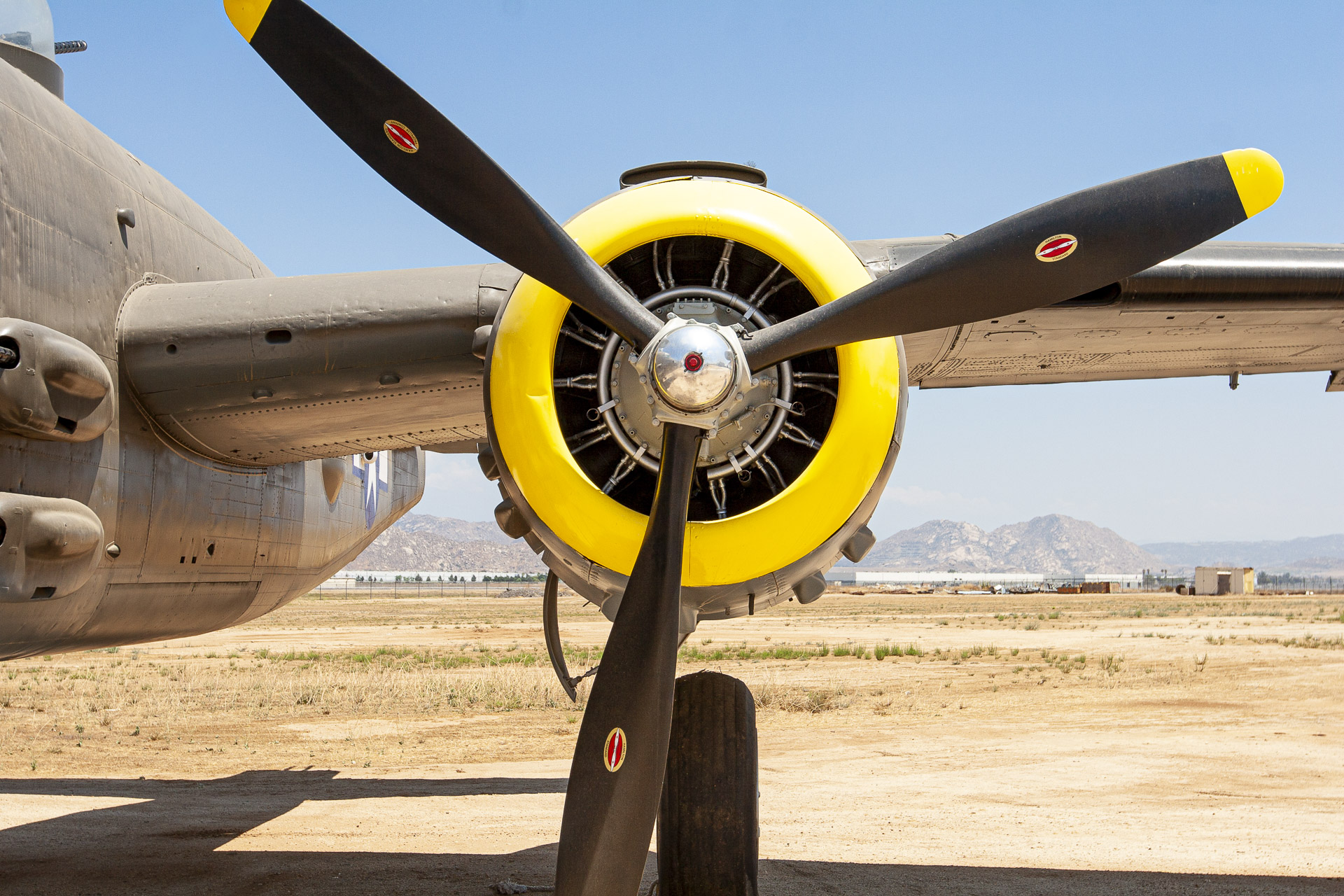 Close-up photograph of a vintage military aircraft's radial engine and three-bladed propeller, featuring a bright yellow engine cowling against a desert landscape.
