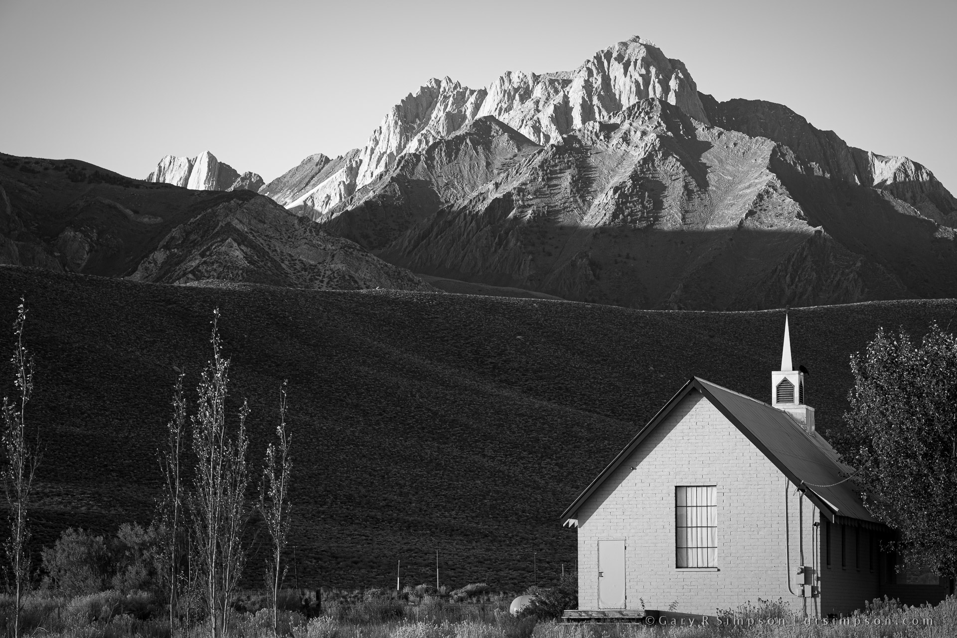 Black-and-white photograph of a small white church with a narrow steeple beneath towering, sunlit Sierra Nevada peaks, with dark desert foothills and sparse vegetation in the foreground.