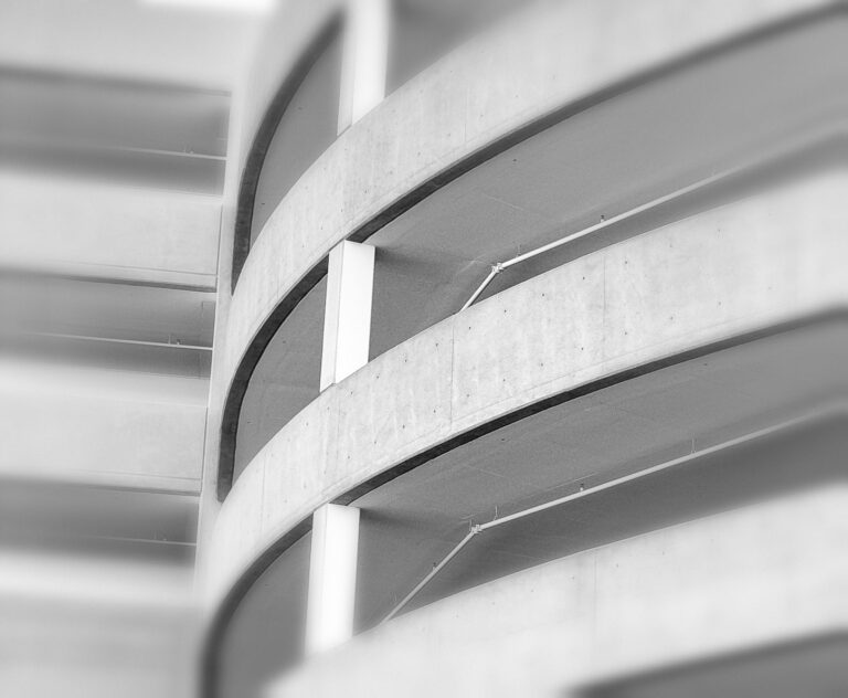 Black-and-white abstract photograph of a multi-level parking structure, featuring sweeping curved concrete ramps, repeating horizontal lines, and softly blurred edges.