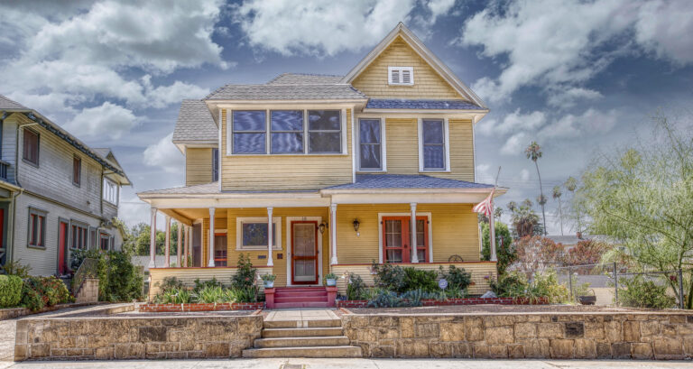 Two-story yellow Victorian house with steep gables, tall windows, a wraparound porch, and stone retaining wall beneath a cloudy sky.