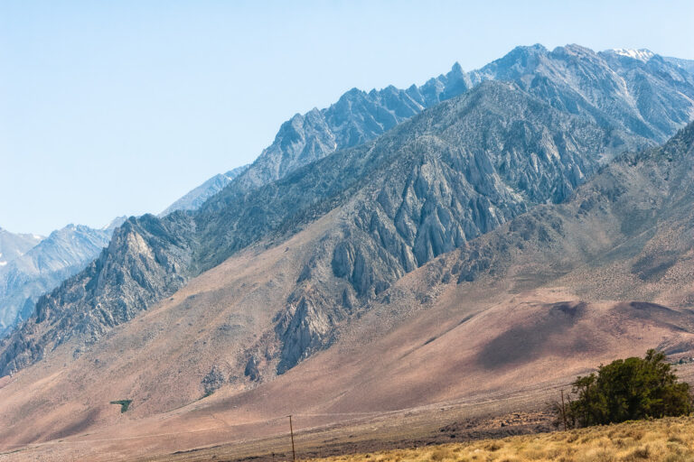 Jagged granite peaks and steep, rocky ridges of the Eastern Sierra Nevada rise above dry, brown foothills along Highway 395, beneath a pale blue sky.