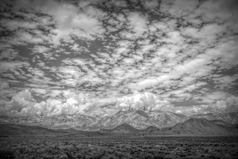 Black-and-white landscape of the Eastern Sierra Nevada, with towering cumulus clouds above rugged mountain peaks and a sky filled with textured, scattered clouds over a sagebrush-covered valley.