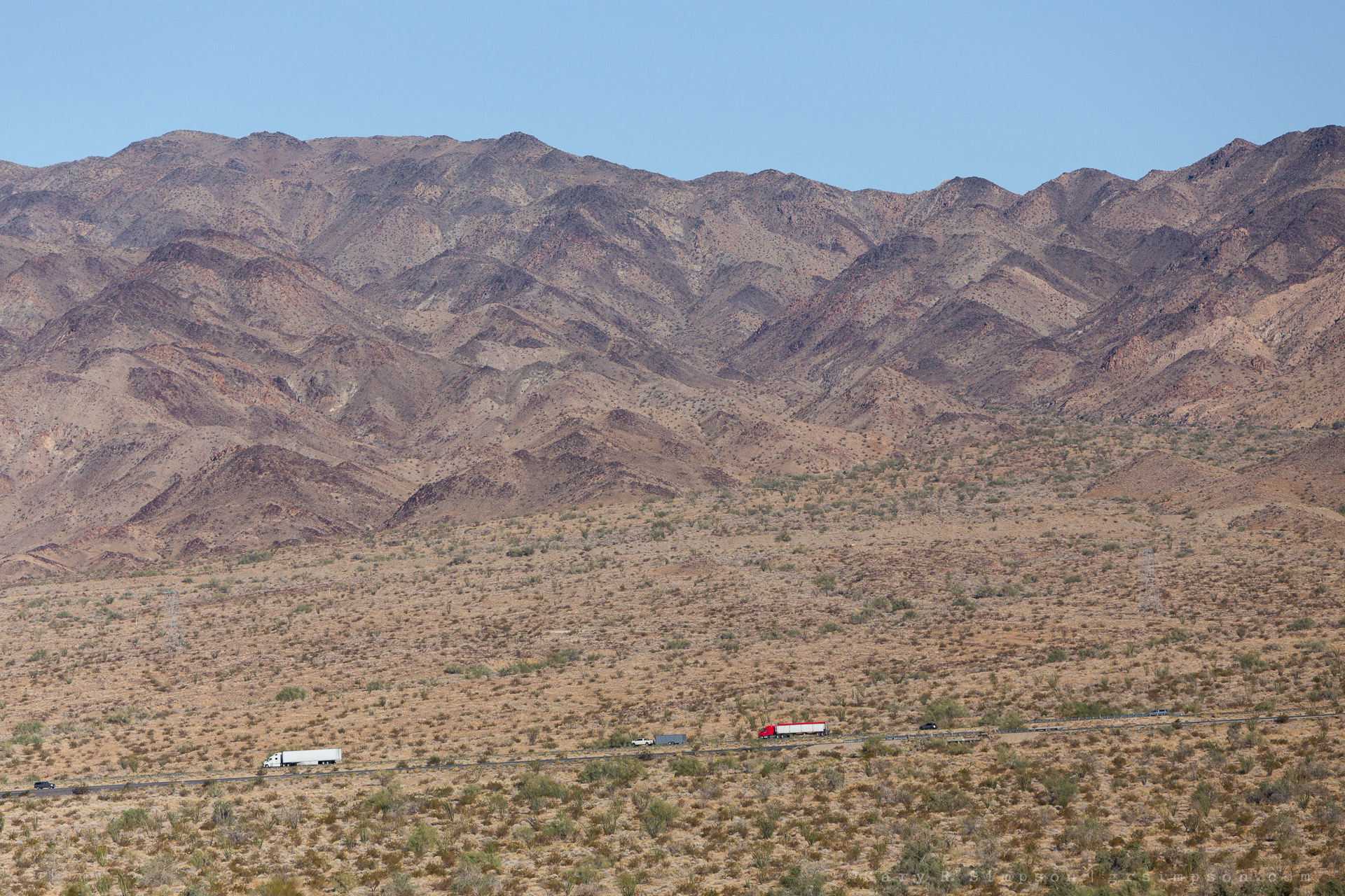 A wide desert landscape with sparse vegetation, trucks traveling along Interstate 10, and rugged mountains rising in the background.
