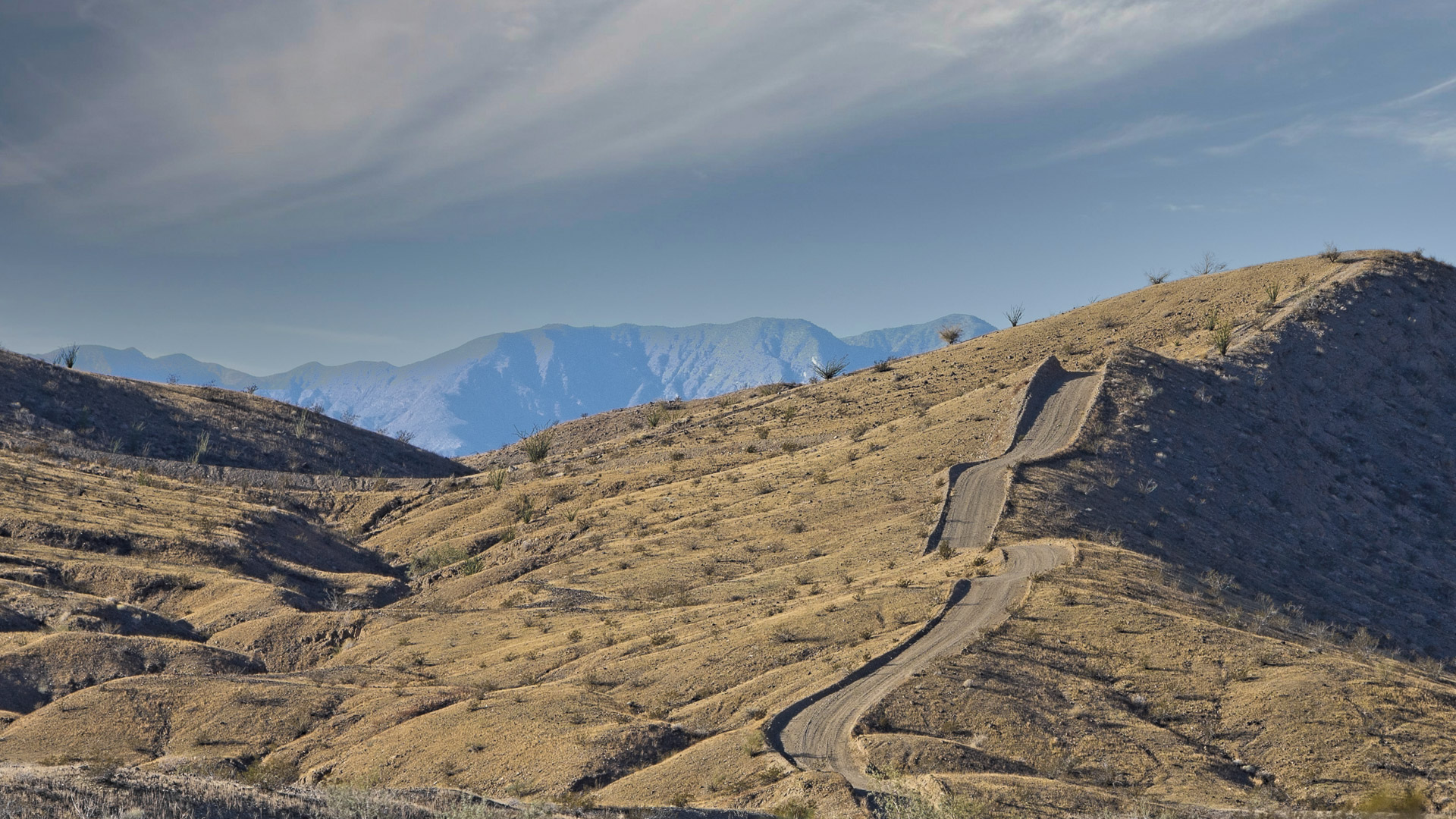 A winding dirt road climbs through barren desert hills toward distant mountains.