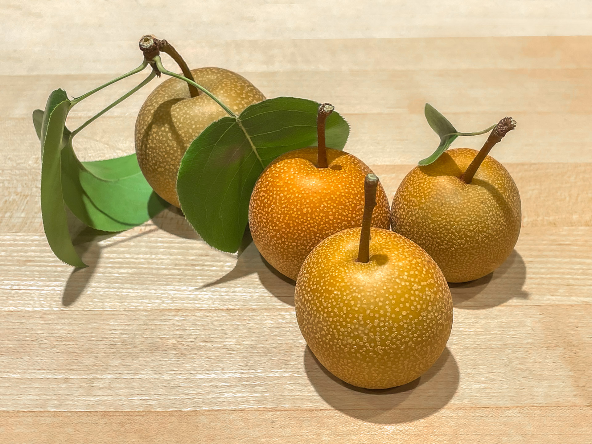 Four golden-brown Asian pears with speckled skins, attached stems, and green leaves arranged on a light wooden surface.