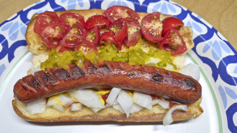 Grilled hot dog in a toasted bun topped with chopped white onions, green relish, mustard, and ketchup, with sliced cherry tomatoes on the upper bun.