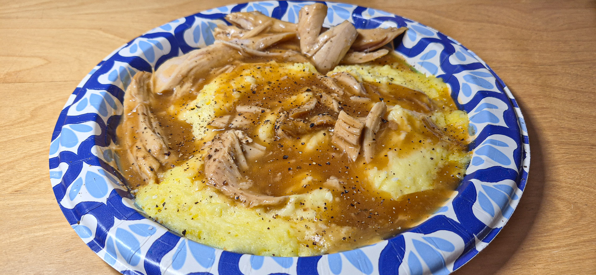 Shredded turkey and brown gravy served over yellow mashed potatoes, sprinkled with black pepper on a blue-and-white patterned paper plate.